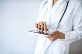 Doctor in a white coat using a digital tablet, with a stethoscope around their neck, focusing on the screen.