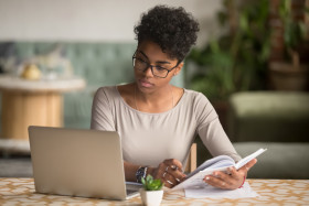 Focused woman wearing glasses works on a laptop while holding a notebook and pen, sitting at a table in a comfortable, home-like environment.