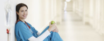 Smiling nurse in blue scrubs sitting in a hospital hallway, holding a green apple and resting against the wall with an IV stand nearby.