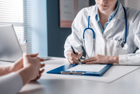Doctor in a white coat with a stethoscope writing on a clipboard during a patient consultation at a desk in a medical office.