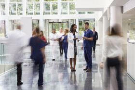 Doctors and medical staff walk through a busy hospital corridor, with two healthcare professionals standing and discussing paperwork in the center.