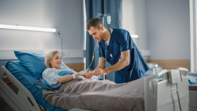 Nurse attending to a smiling female patient lying in a hospital bed in a bright, modern hospital room.