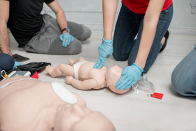 Person wearing blue gloves performing infant CPR on a baby mannequin during a first aid training session, with CPR equipment visible nearby.