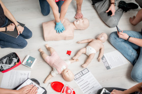 People in a first aid training session practicing CPR on adult and infant mannequins, surrounded by medical kits, first aid instructions, and notepads on the floor.