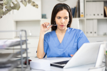 Female healthcare professional in blue scrubs sitting at a desk, looking thoughtfully at a laptop screen while holding a pen to her lips in an office setting.