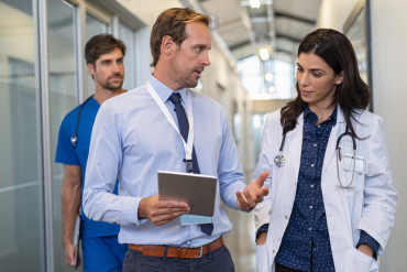 Medical professionals and an administrator discuss in a hospital hallway, with one holding a tablet and a doctor wearing a white coat and stethoscope.