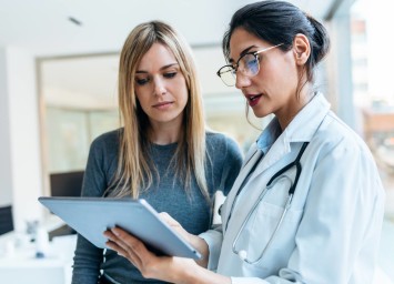 Female doctor with stethoscope showing information on a tablet to a female patient in a bright medical office.