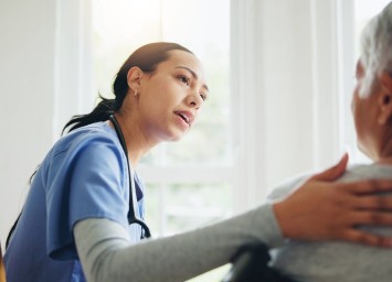 Nurse in blue scrubs comforting an elderly patient, gently touching their shoulder while speaking empathetically in a bright, sunlit room.
