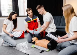 Four people practicing CPR on a training manikin, with two holding instruction sheets and two using a resuscitation bag, in a classroom setting.