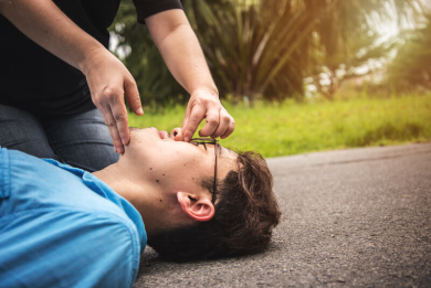 Person performing rescue breathing, pinching the nose of an unconscious man lying on the ground outdoors, preparing to give mouth-to-mouth resuscitation.