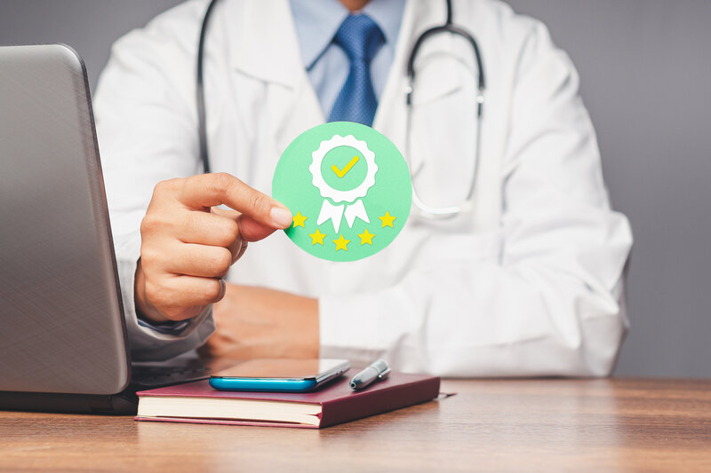 a desk with books, notepad and pen on the table with a close up of a doctor’s hand holding up a green badge that means completion