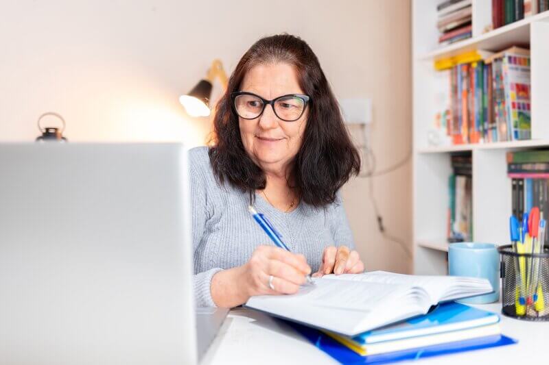 an older adult female wearing glasses sits on a desk and looking at a laptop screen while holding a pen studies online with CEU credits