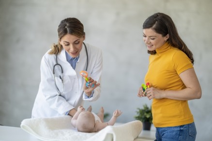A PALS certified female doctor is holding up a rattle in front an infant lying on a clinic bed with the mother beside them