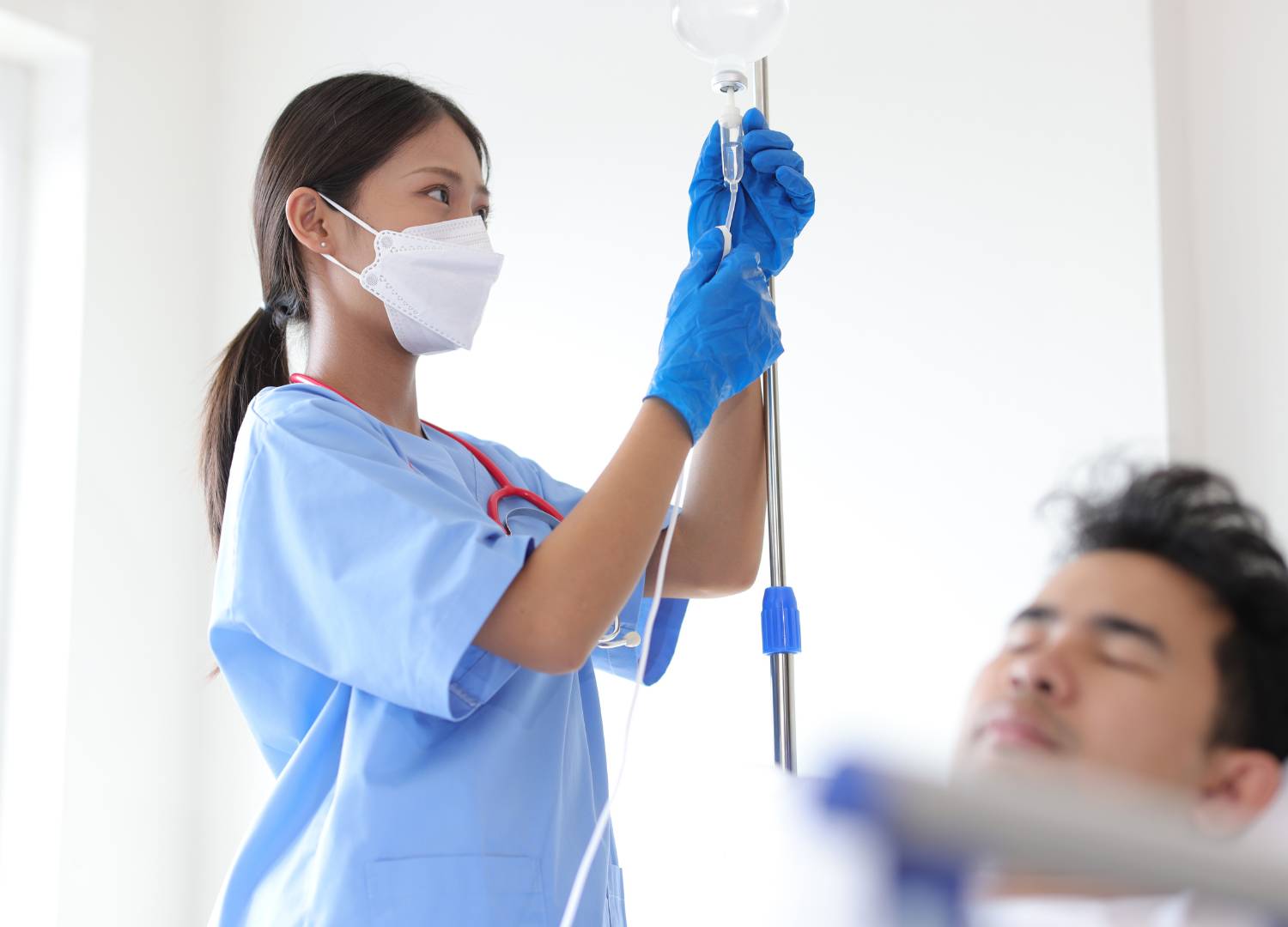 A nurse wearing a mask and gloves administers an IV to a patient.